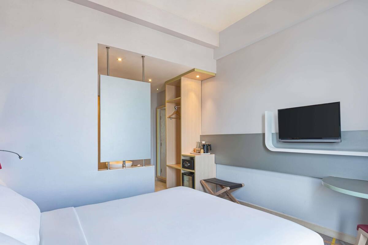 A hotel room with a bed, a TV mounted on the wall, a desk with a chair, and a mirror.