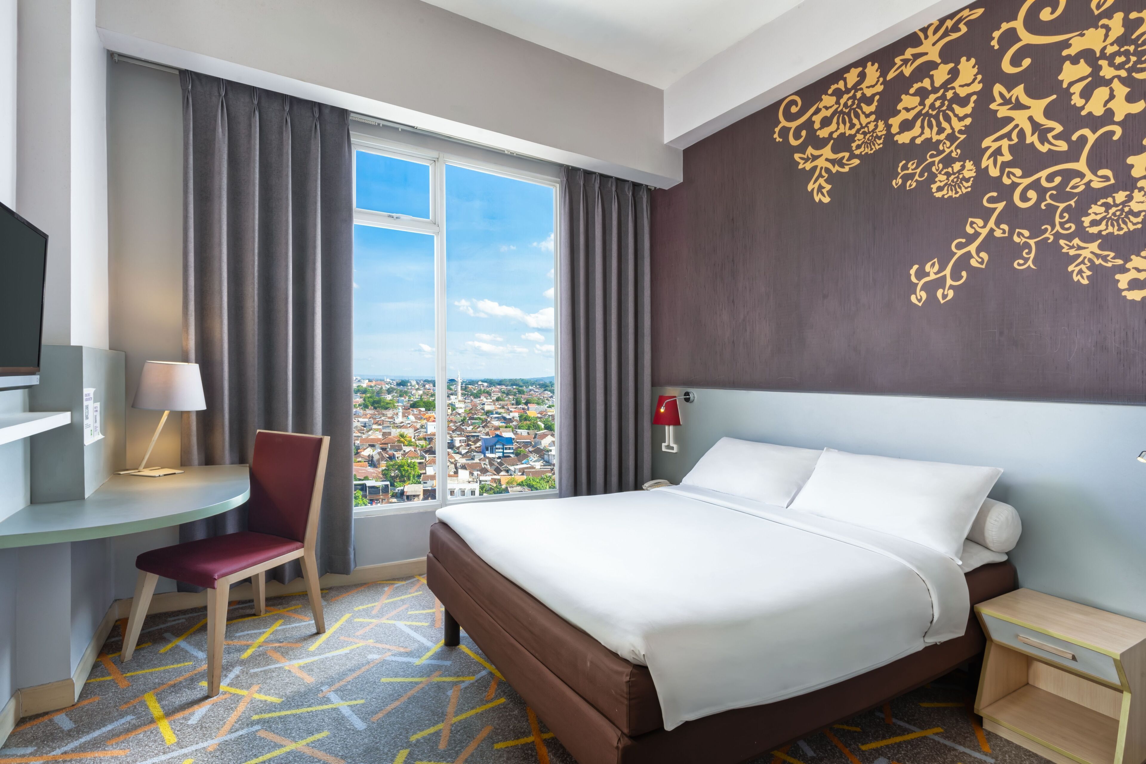 A hotel room with a large bed, a desk with a chair, a TV, a window with curtains, and a decorative wall.
