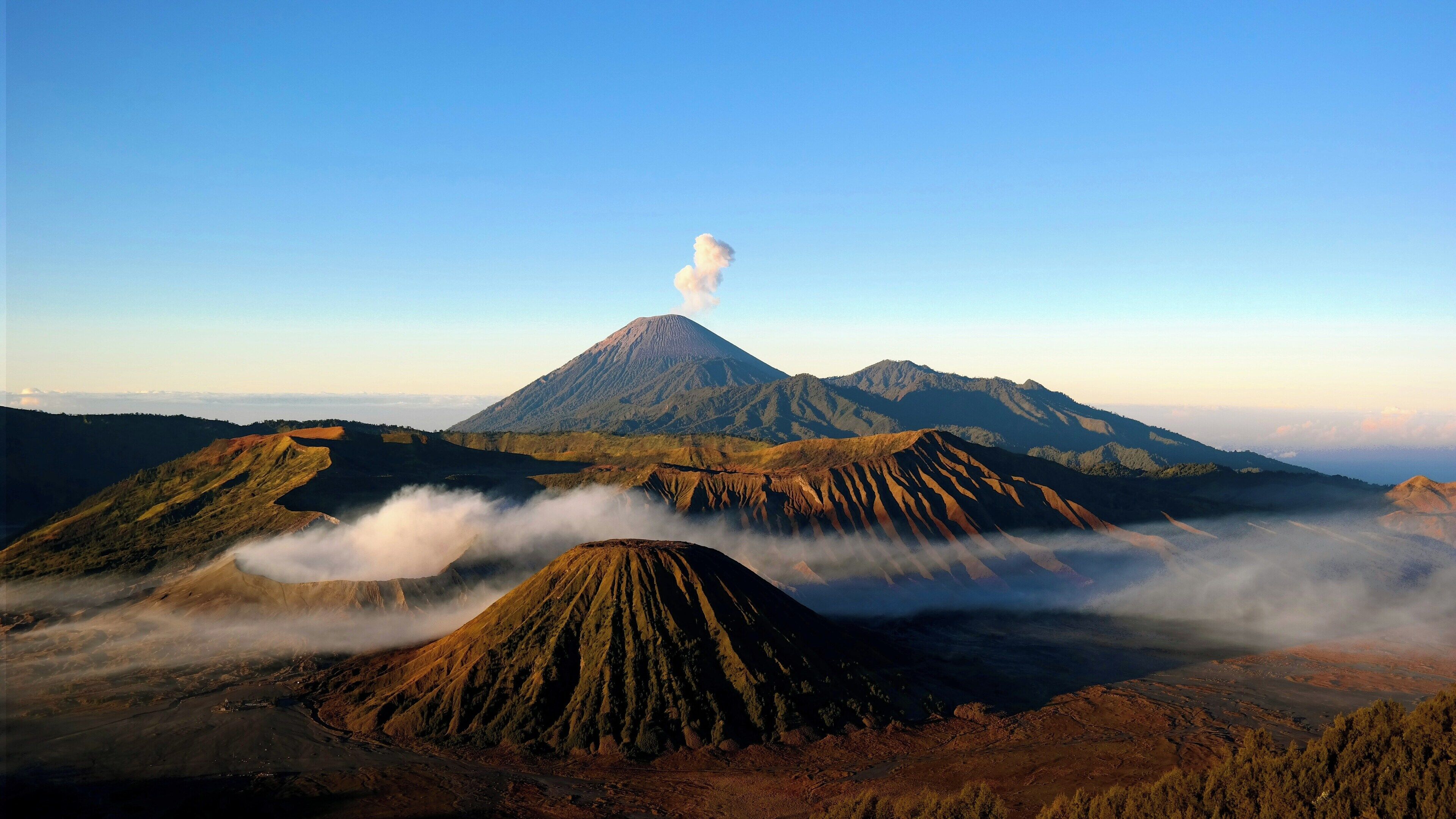 Un paisaje volcánico con un pico montañoso destacado que emite humo, rodeado de colinas y valles cubiertos de bosque.