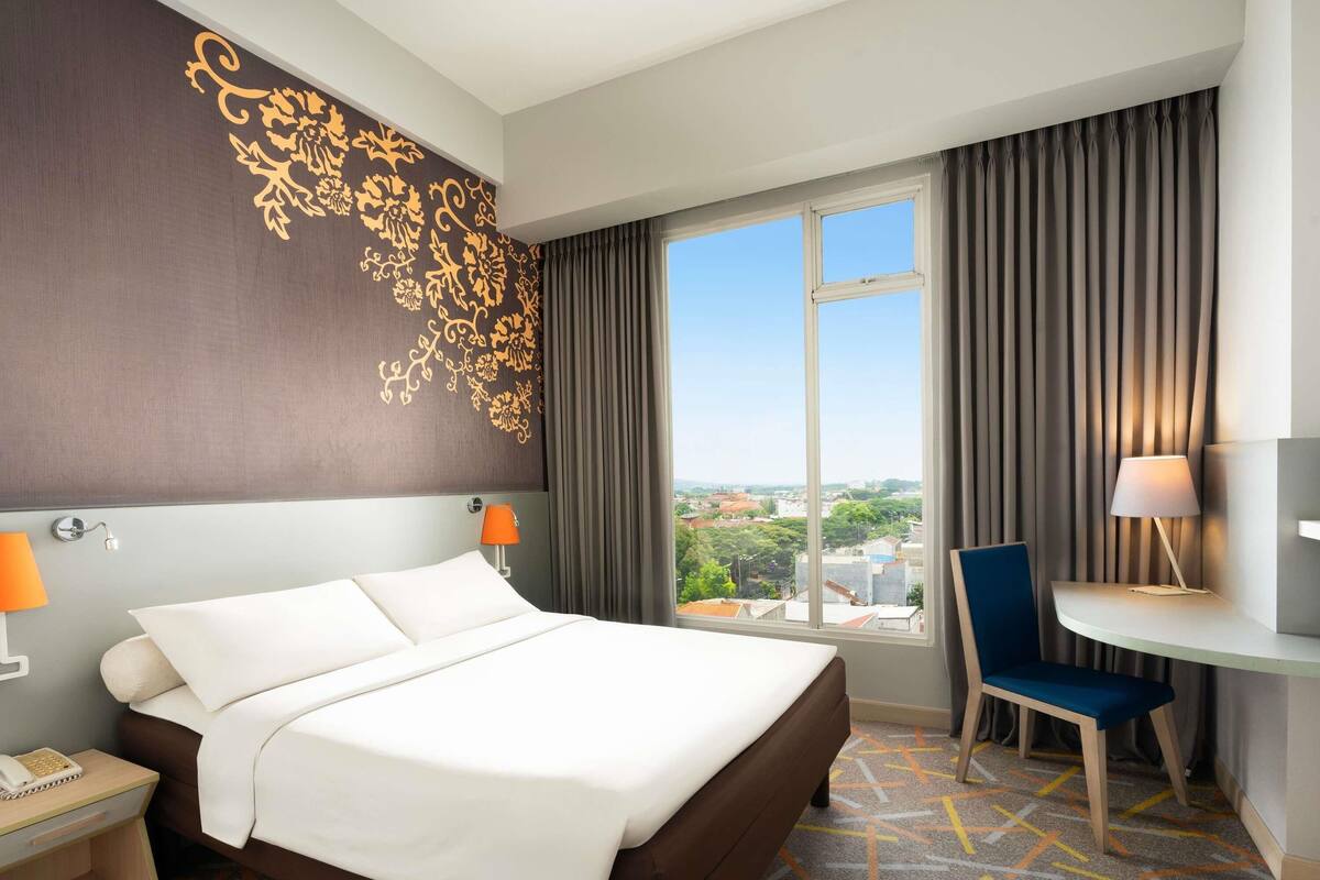 A hotel room with a large bed, a desk with a chair, a view of the city, and a decorative wall.