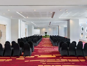 Une salle de conférence avec des rangées de chaises, un écran de projection et un sol recouvert de moquette.