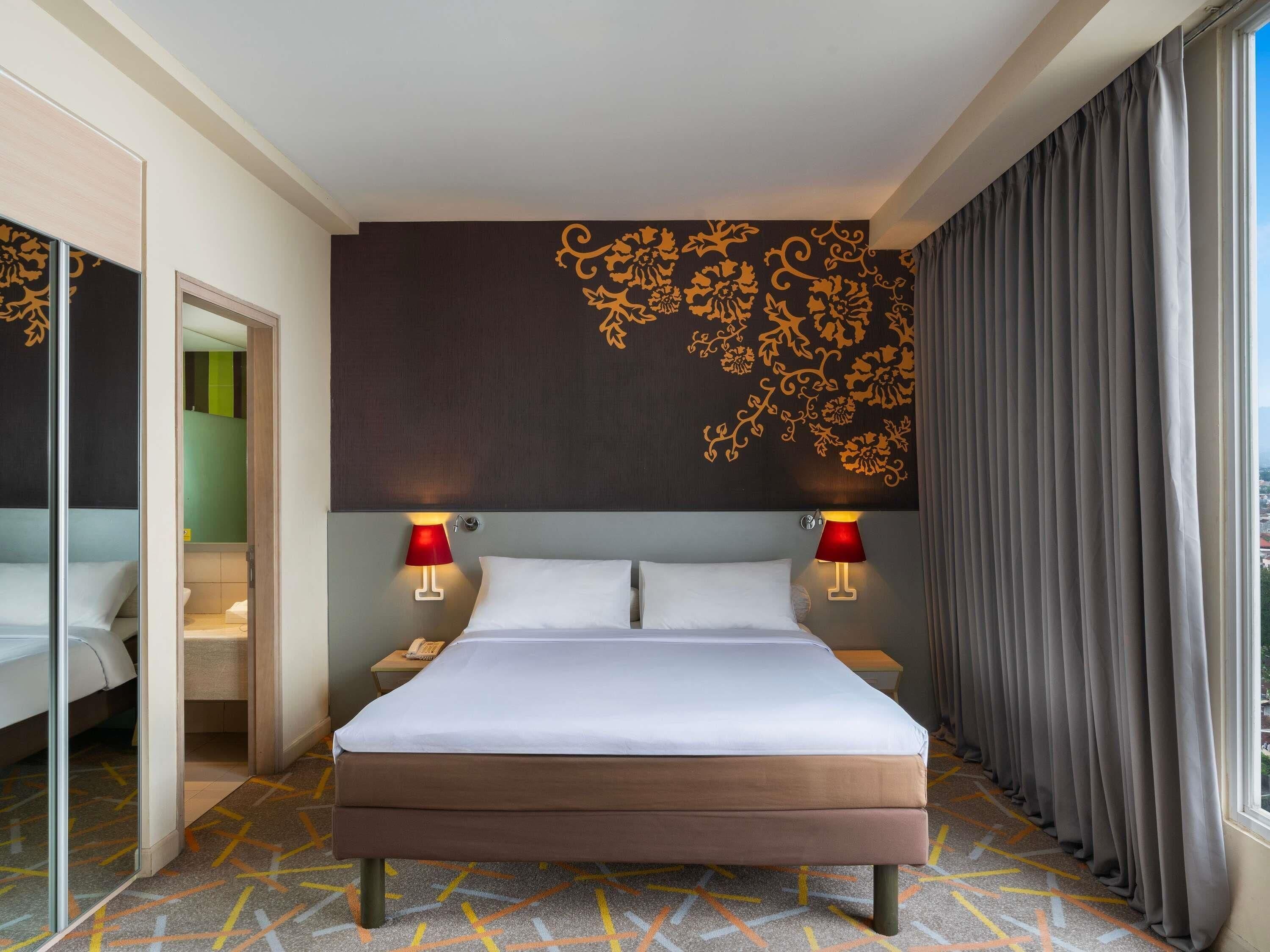 A hotel room with a large bed, two bedside lamps, a wall with floral patterns, a window with curtains, and a patterned carpet.