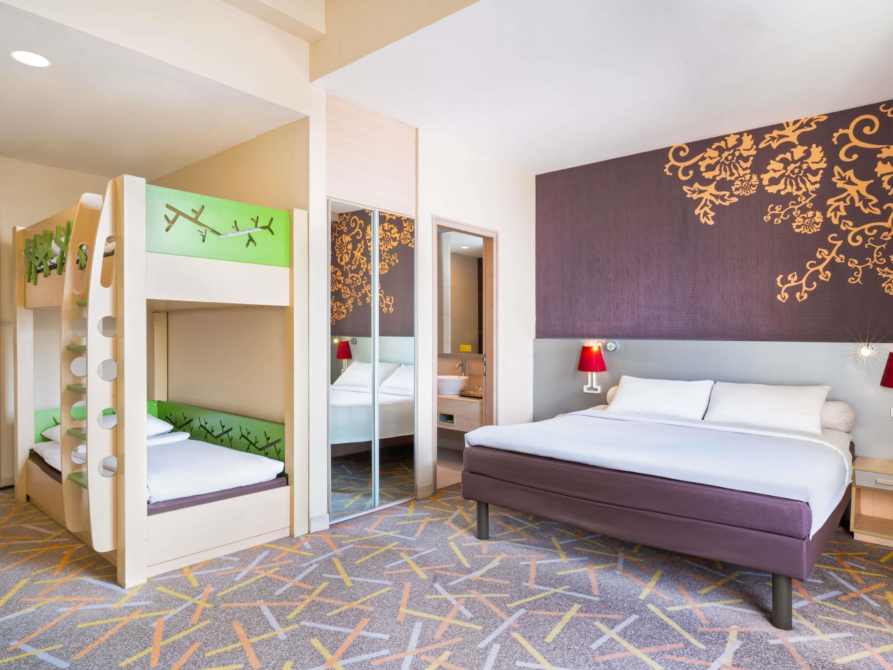 A hotel room with a bunk bed, a single bed, a mirror, a bathroom with a sink, and a patterned carpet.