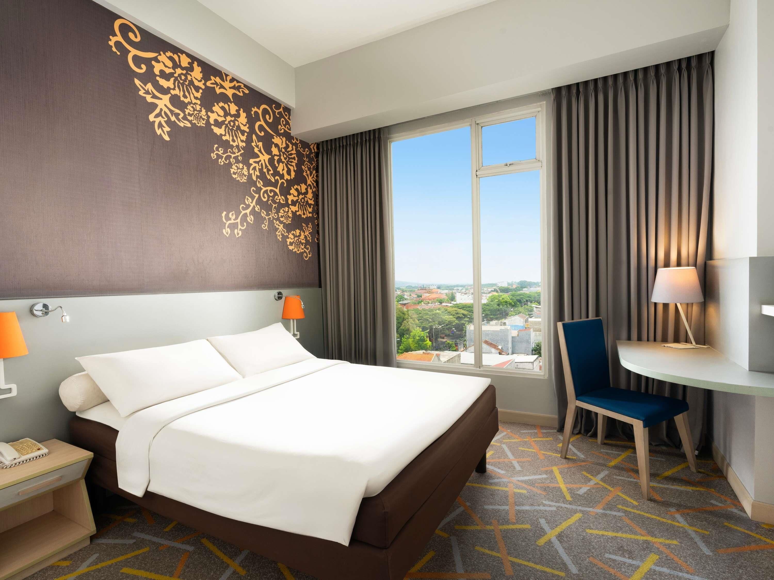 A hotel room with a large bed, a desk with a chair, a view of the city, and a decorative wall.