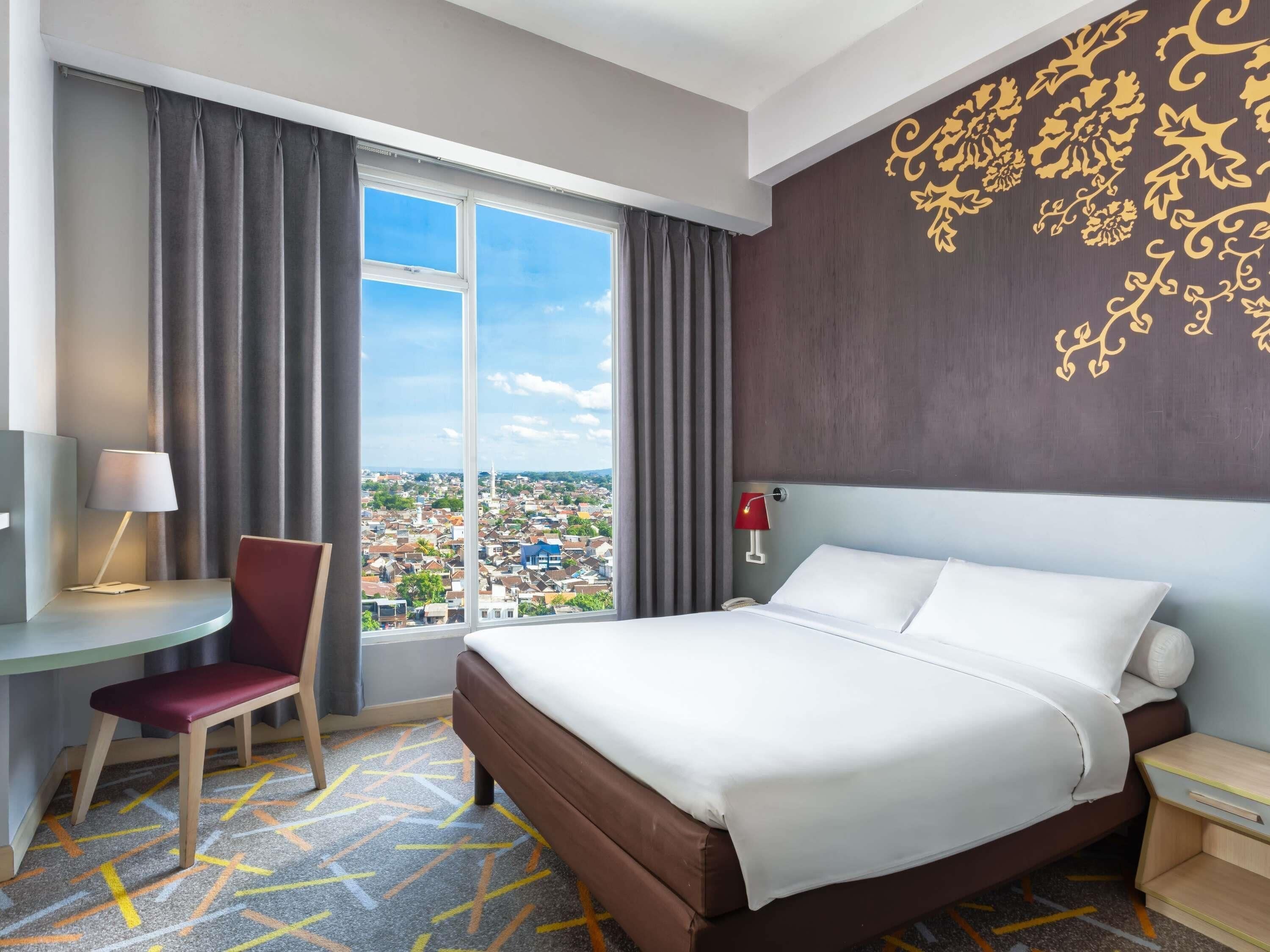 Habitación de hotel con una cama grande, un escritorio con silla, una mesita de noche y vistas a la ciudad a través de un ventanal amplio.