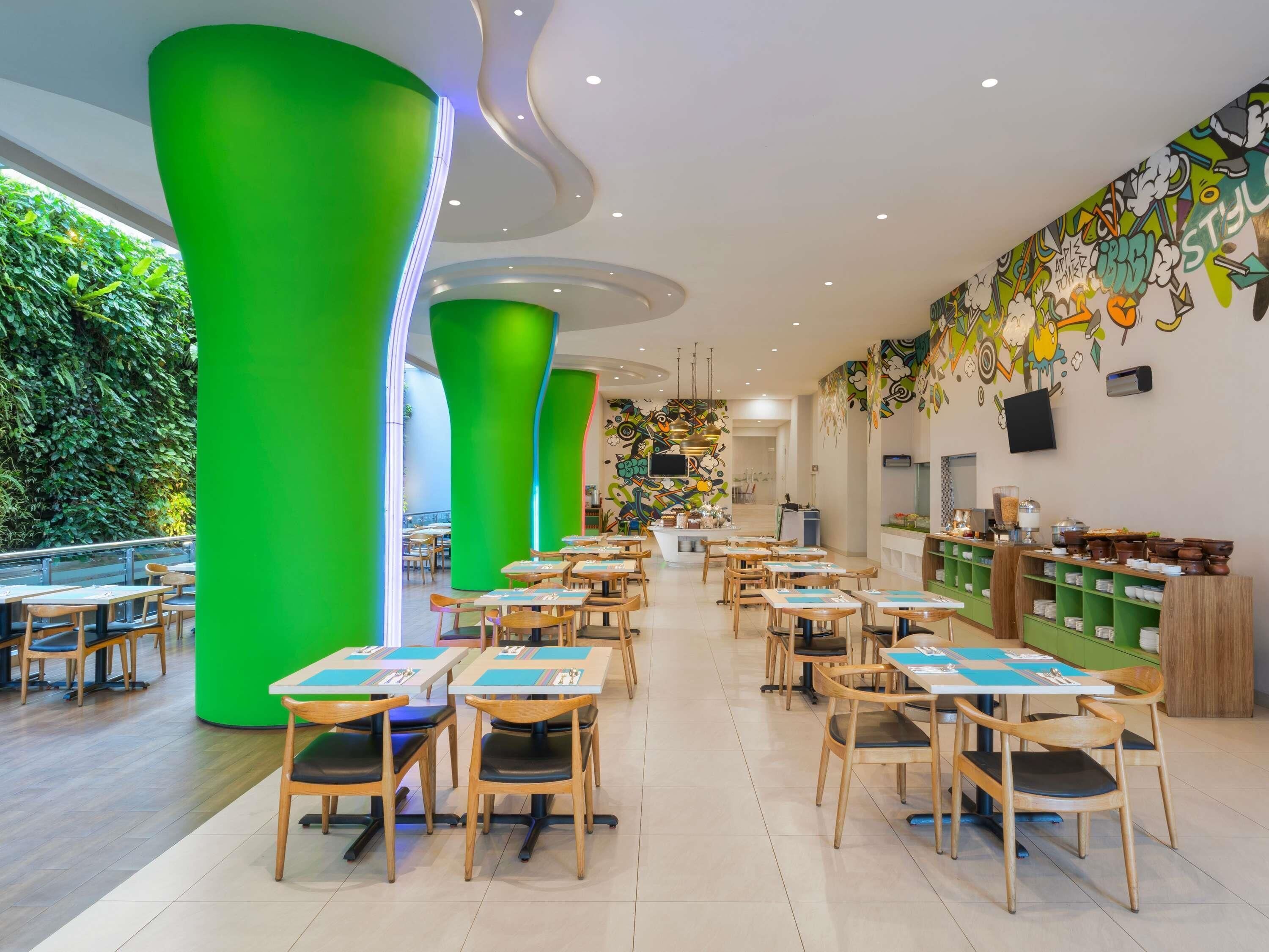 A modern restaurant with green pillars, wooden tables, and a colorful mural on the wall.