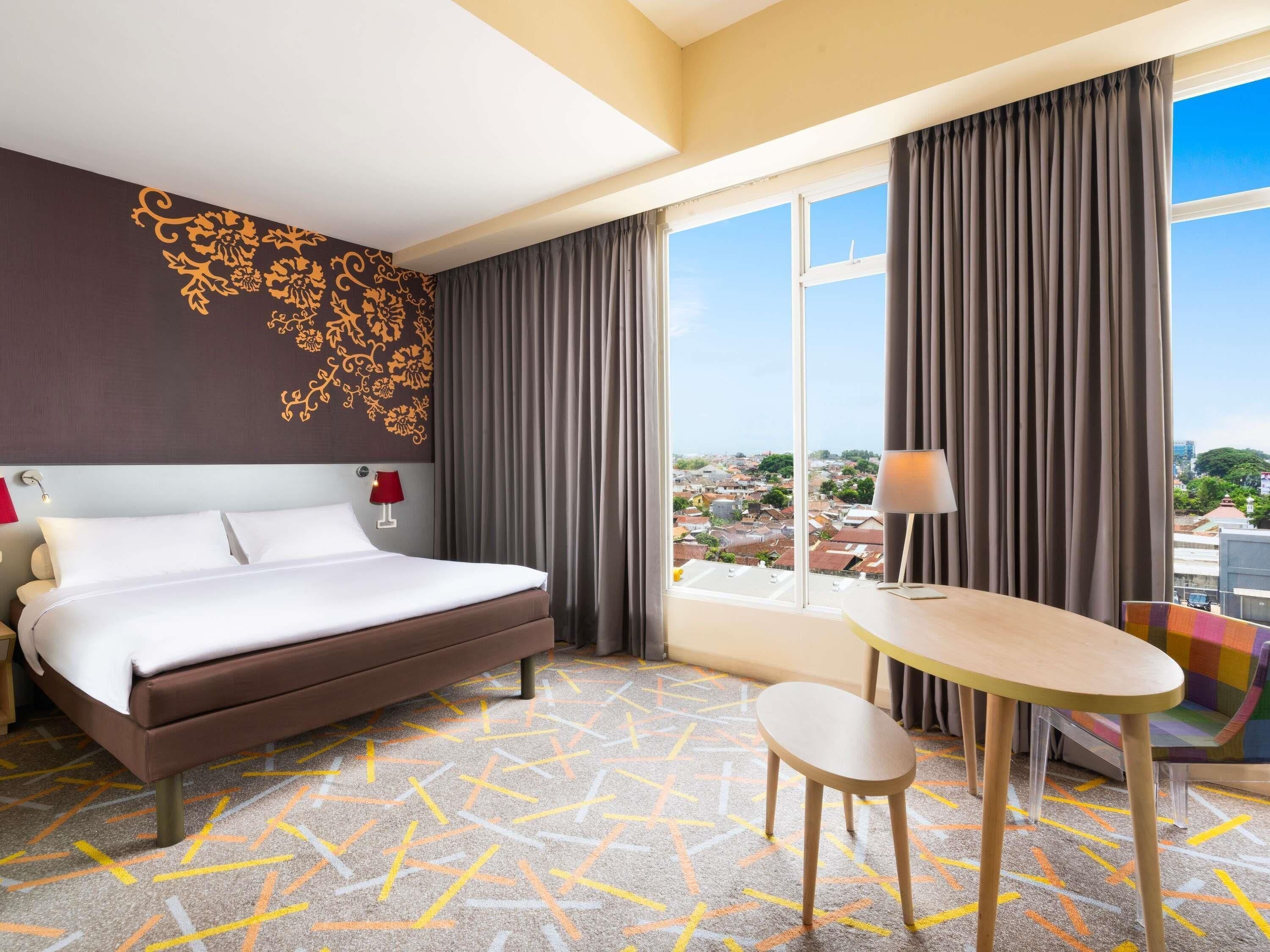 A hotel room with a bed, a desk, a chair, a view of the city, and a patterned carpet.
