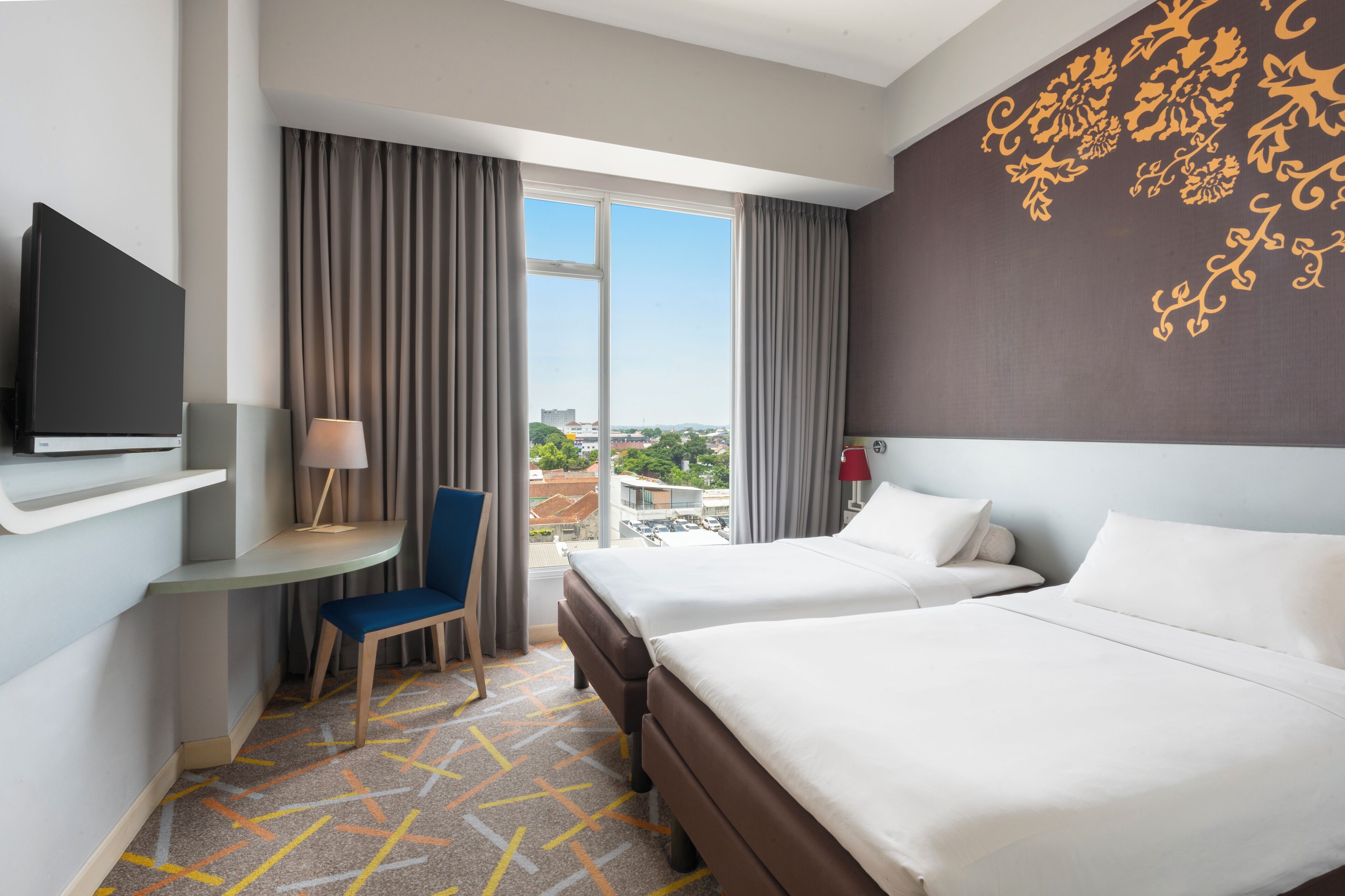 Une chambre d’hôtel avec deux lits, un bureau, une chaise, une télévision et une vue sur la ville grâce à de grandes fenêtres.