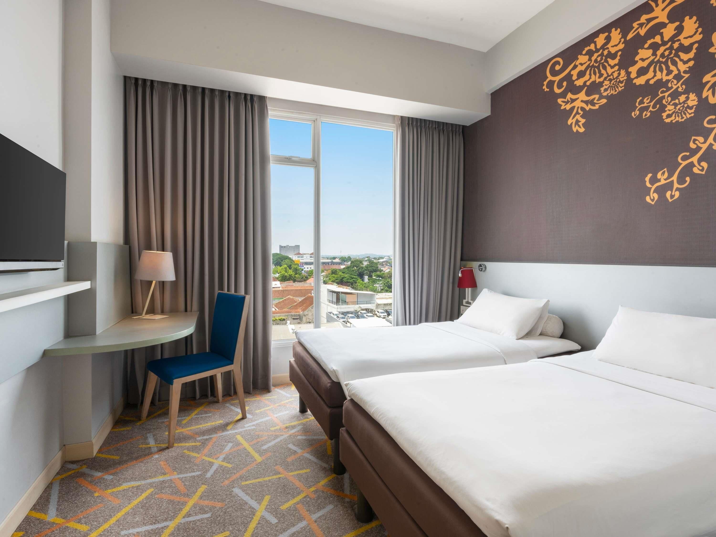 A hotel room with two beds, a desk, a chair, a TV, and a large window with curtains.