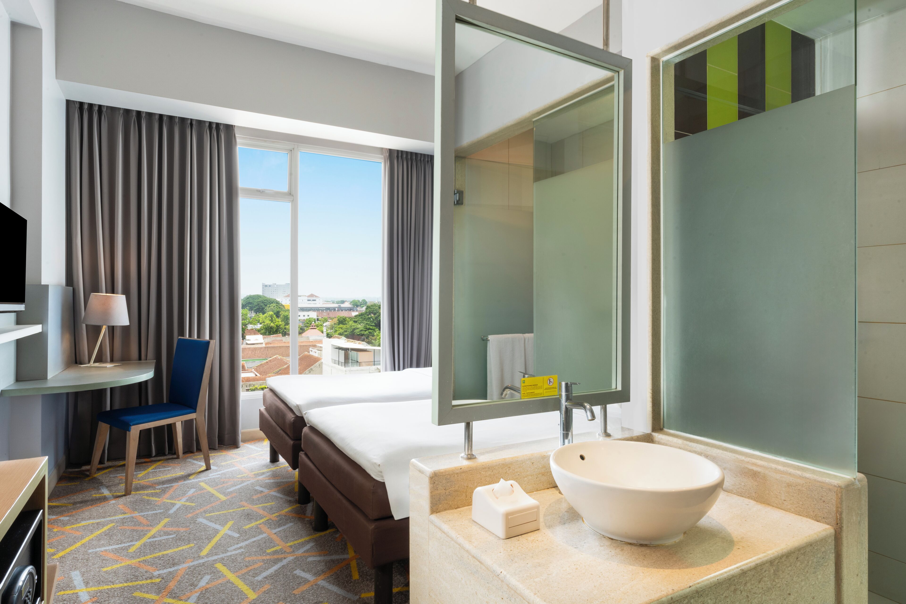 A hotel room with a large bed, a desk with a chair, a bathroom with a large mirror, and a view of the city.
