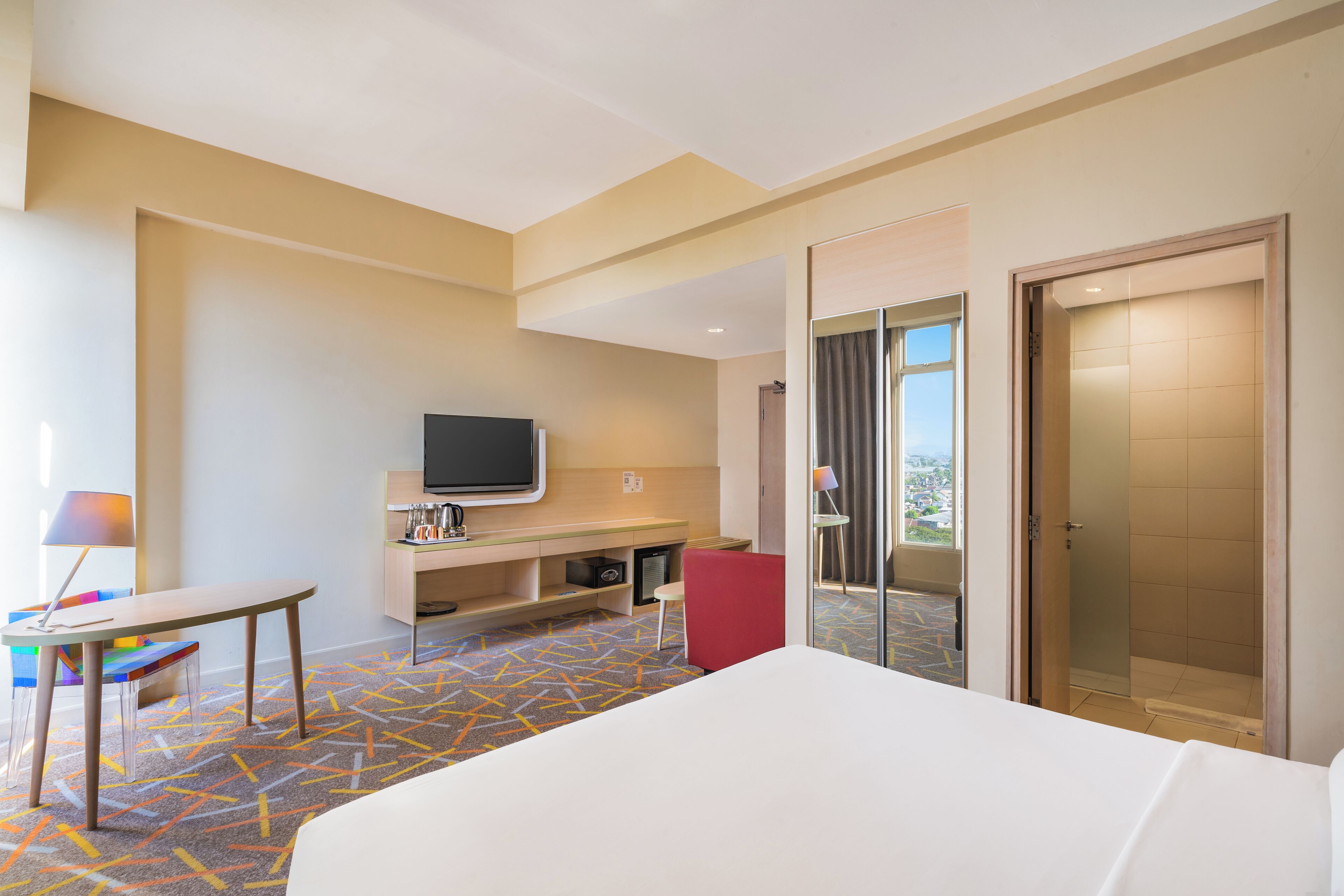 A hotel room with a bed, a desk, a television, a shower, and a view of the city.