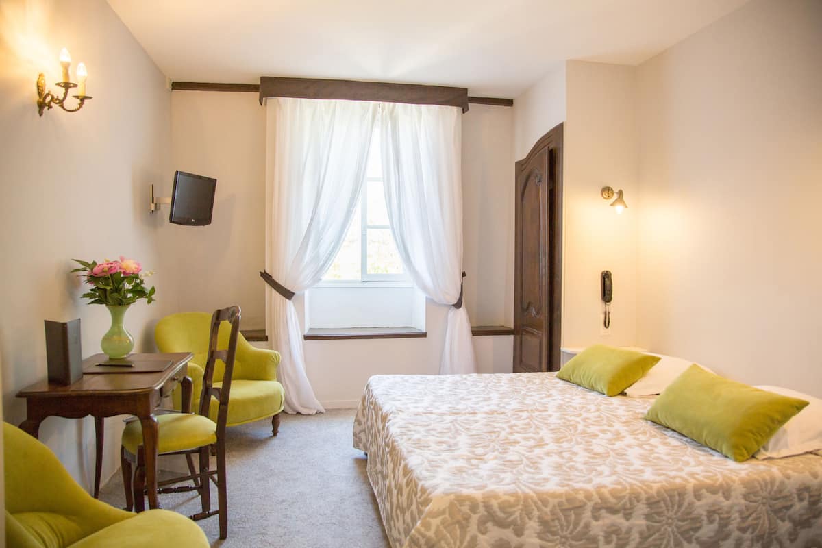 A hotel room with a bed, two chairs, a small table with a vase, a TV mounted on the wall, and a window with curtains.