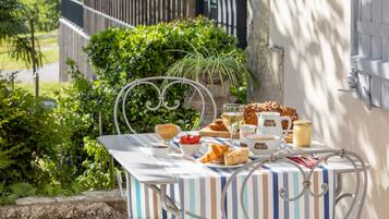 Un área de comedor al aire libre con una mesa preparada para una comida, rodeada de follaje y un edificio al fondo.