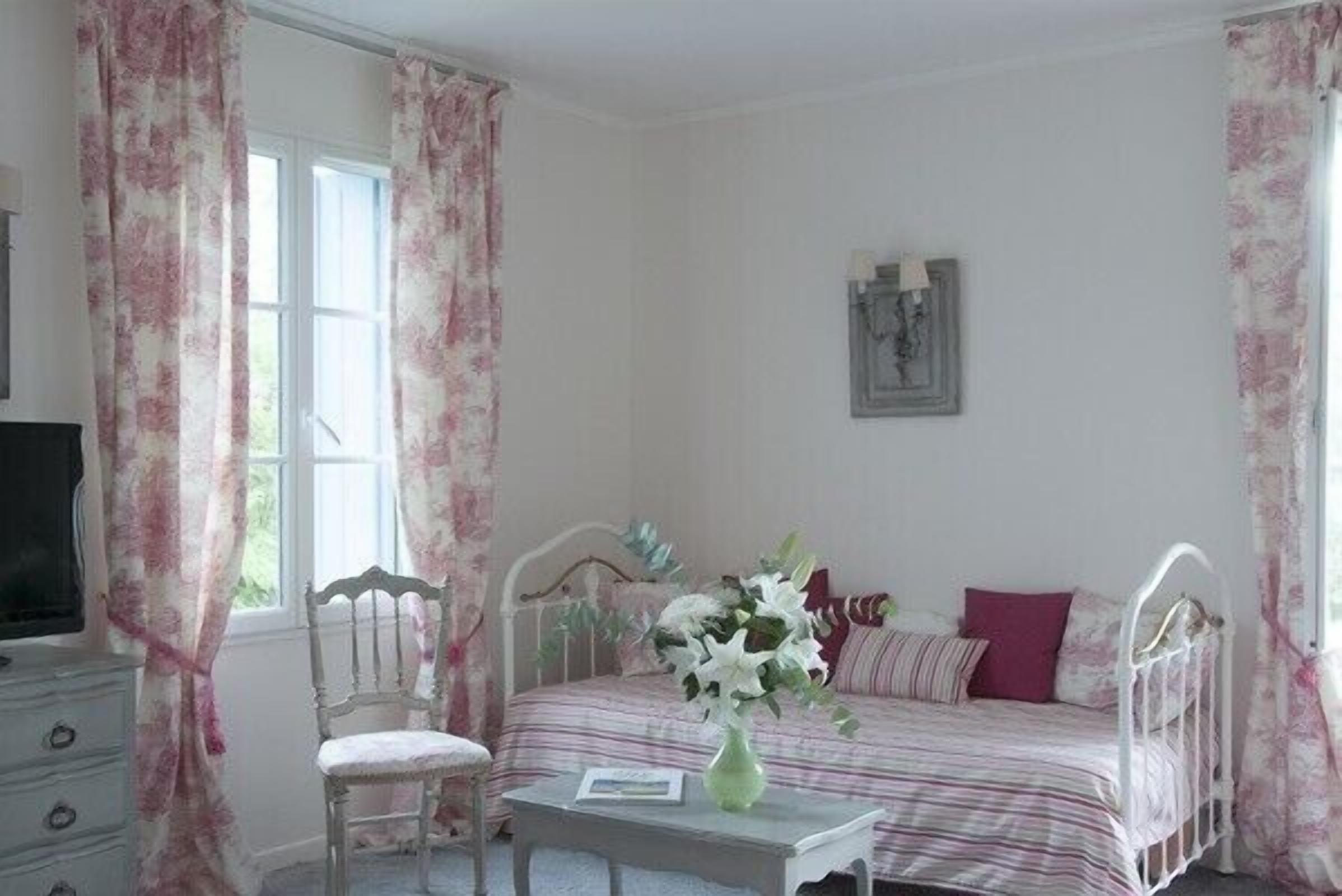 Une chambre à coucher avec un lit, une chaise, une table avec un vase et des rideaux.