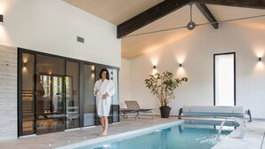 A modern indoor pool area with a glass door entrance, a lounge chair, and a potted plant. - Château Bellevue (Cazaubon)