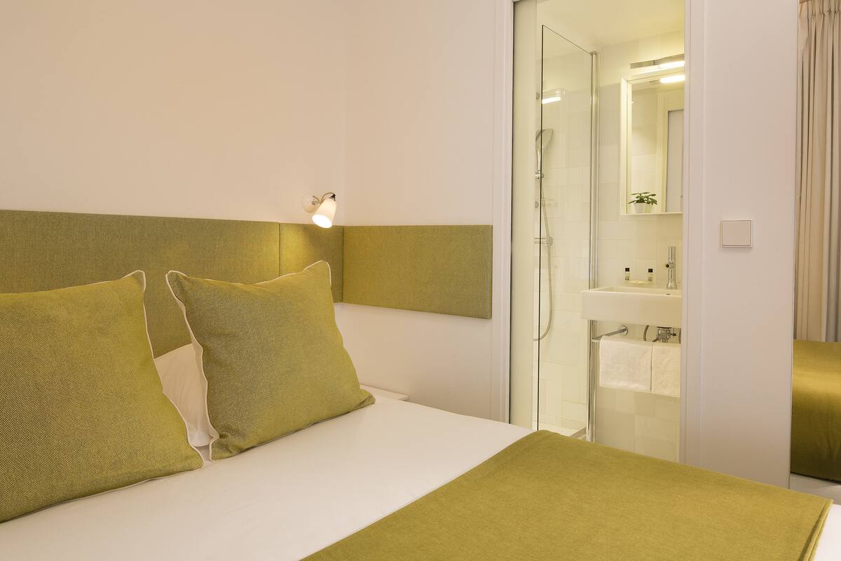 Habitación de hotel con cama, mesita de noche, espejo, zona de ducha y baño con lavabo y toallero.