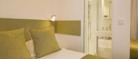Habitación de hotel con cama, mesita de noche, espejo, zona de ducha y baño con lavabo y toallero.