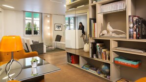 A modern living room with a glass coffee table, orange chairs, a bookshelf with books, a globe, and a decorative bird sculpture. - Hôtel Mistral (Paris)