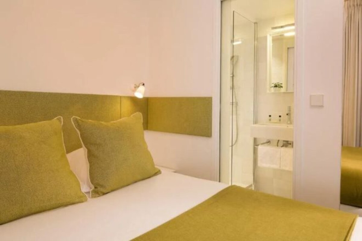 A hotel room with a bed, a bedside table, a nightstand, a mirror, and a bathroom visible through an open door.