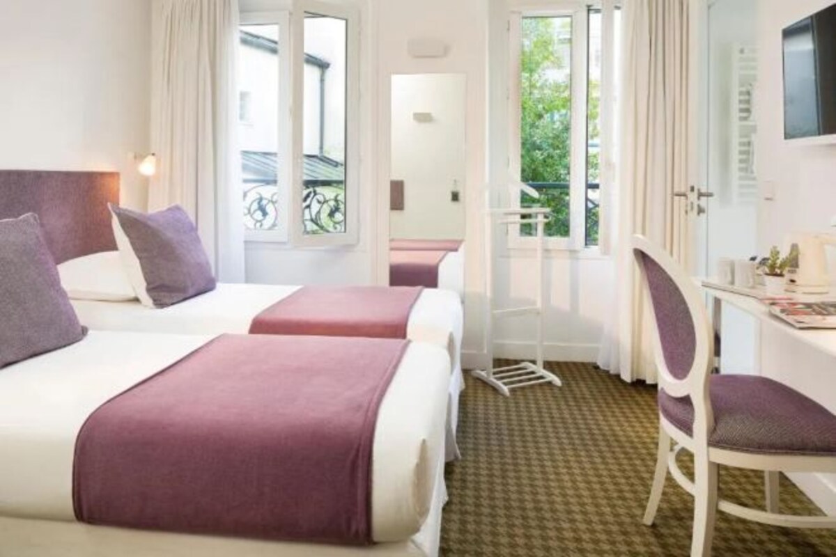 A hotel room with two beds, a desk, and a chair. There is a window with curtains and a view of greenery outside.