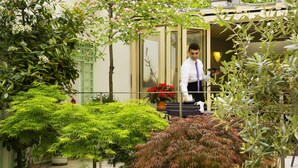A hotel entrance with a glass door, a potted plant with red flowers, and lush greenery. - Hôtel Mistral (Paris)