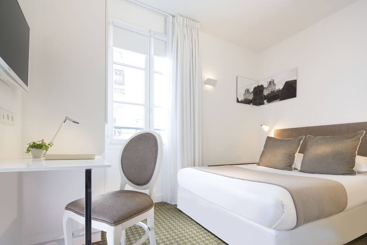 Habitación de hotel con cama, escritorio, silla, ventana con cortinas e imagen colgada en la pared.