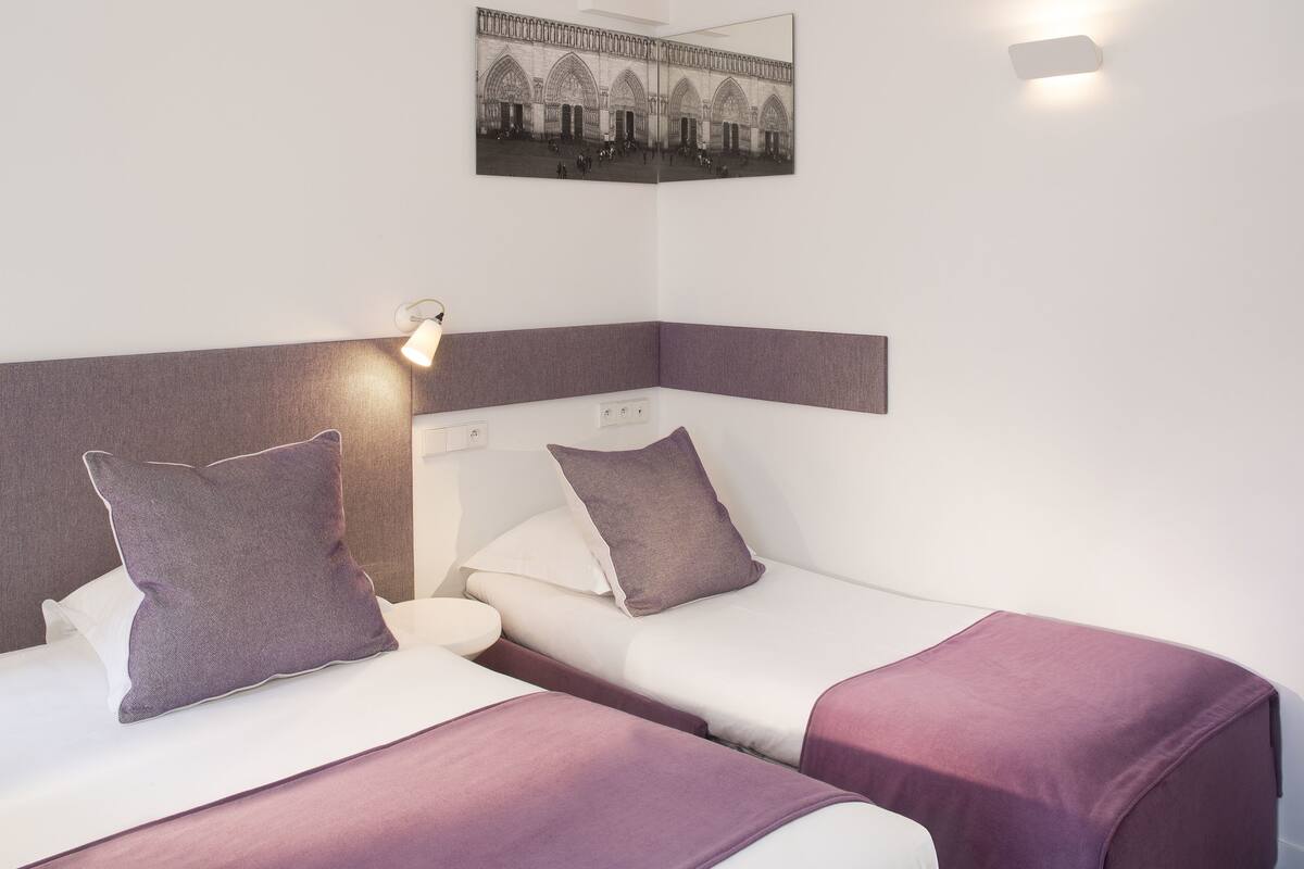 Habitación de hotel con dos camas, cada una con cubrecama y almohadas moradas a juego. Se observa un cuadro colgado en la pared que representa una catedral, una lámpara de noche y una mesita de noche pequeña.