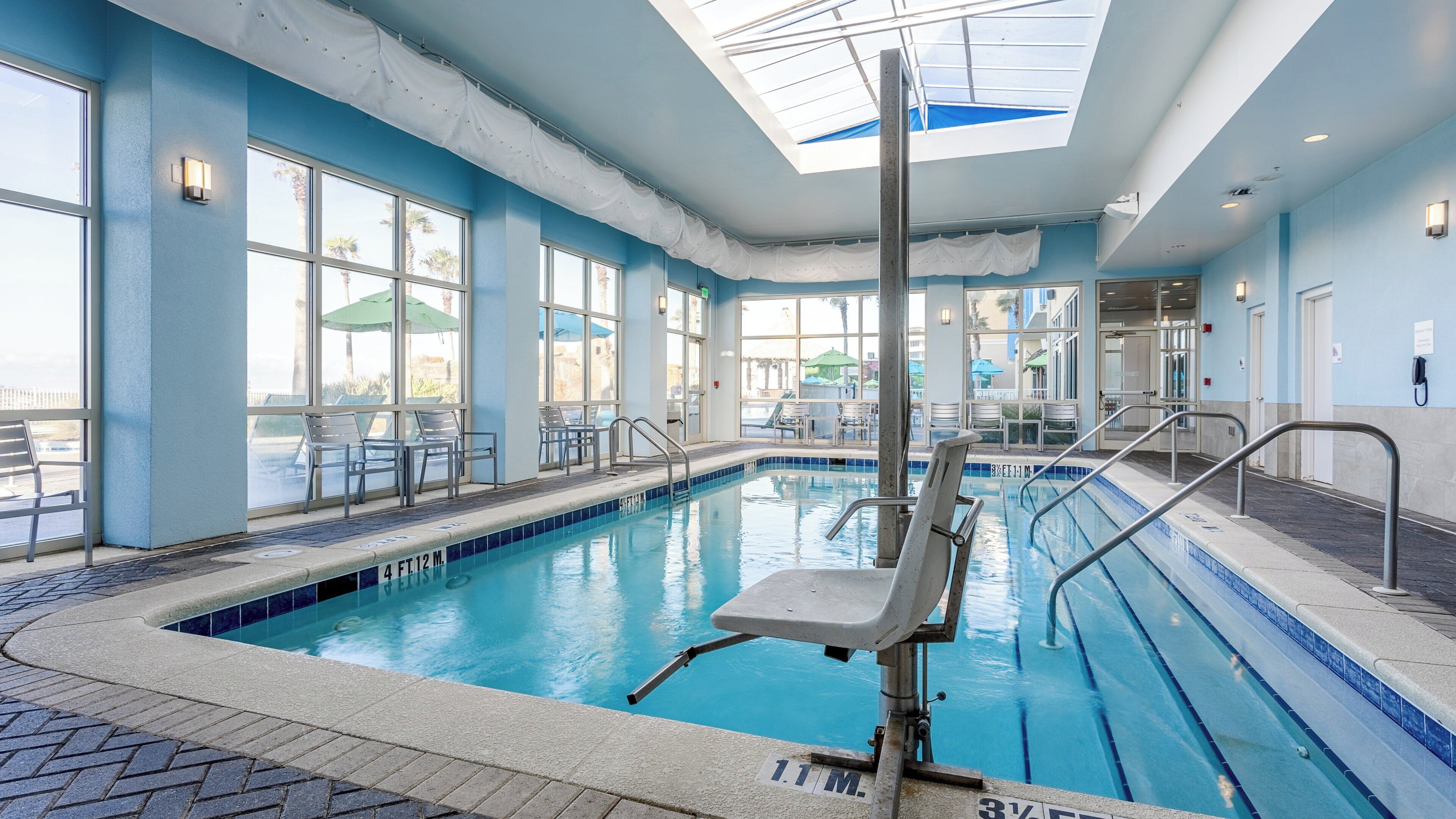 An indoor swimming pool with clear blue water, surrounded by windows and chairs.