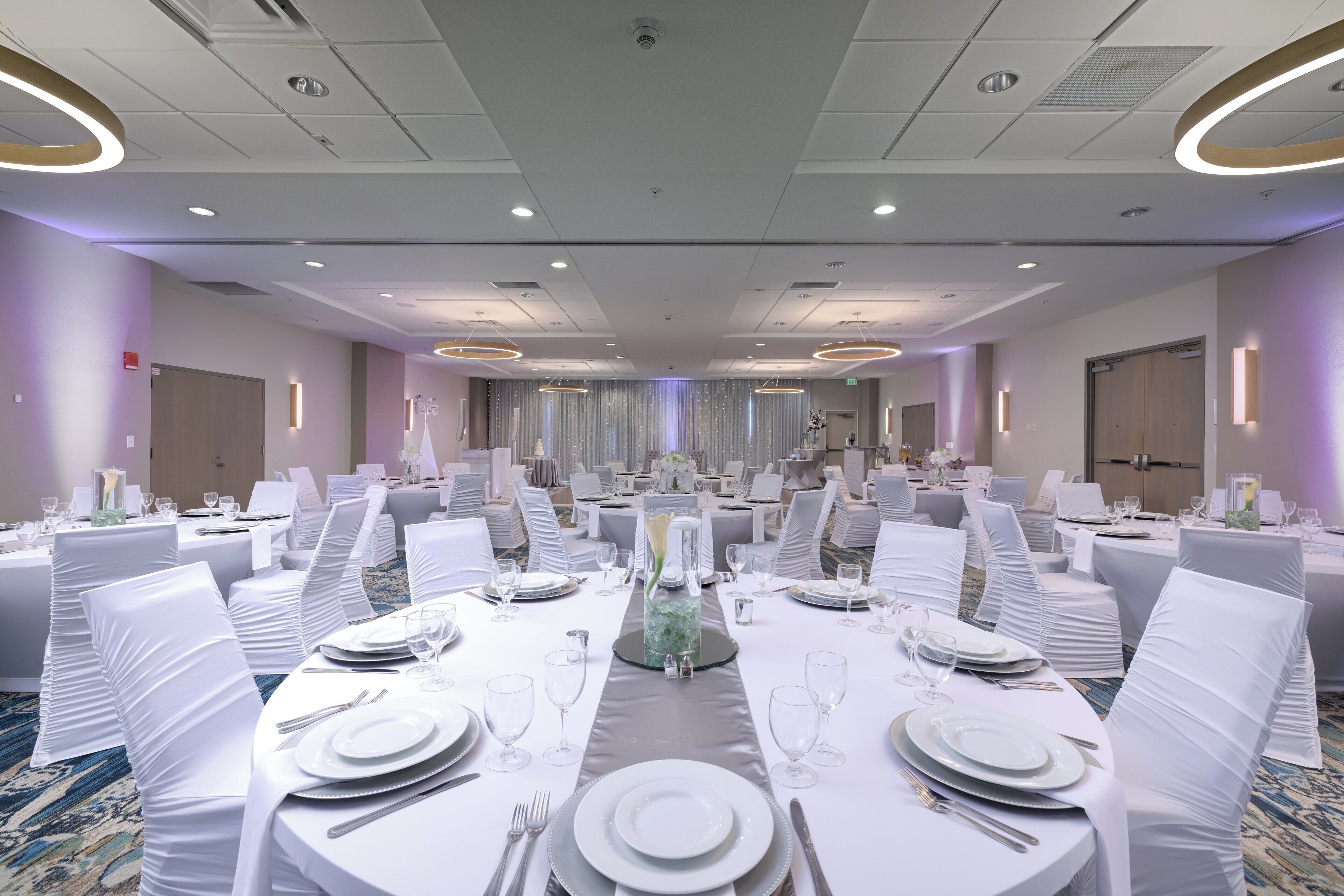 A banquet hall with round tables set for dining, white tablecloths, and chairs with white covers.