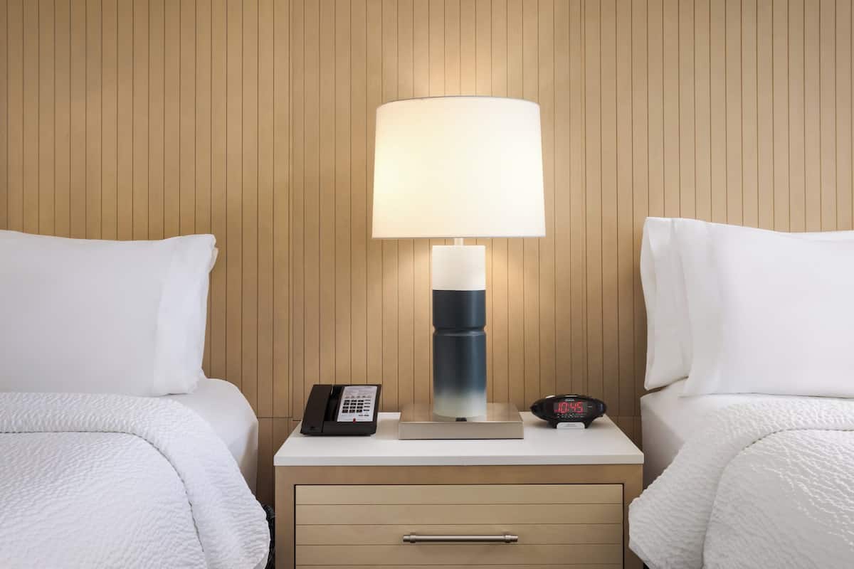 A hotel room with two beds, a nightstand with a lamp, a phone, and a digital clock.
