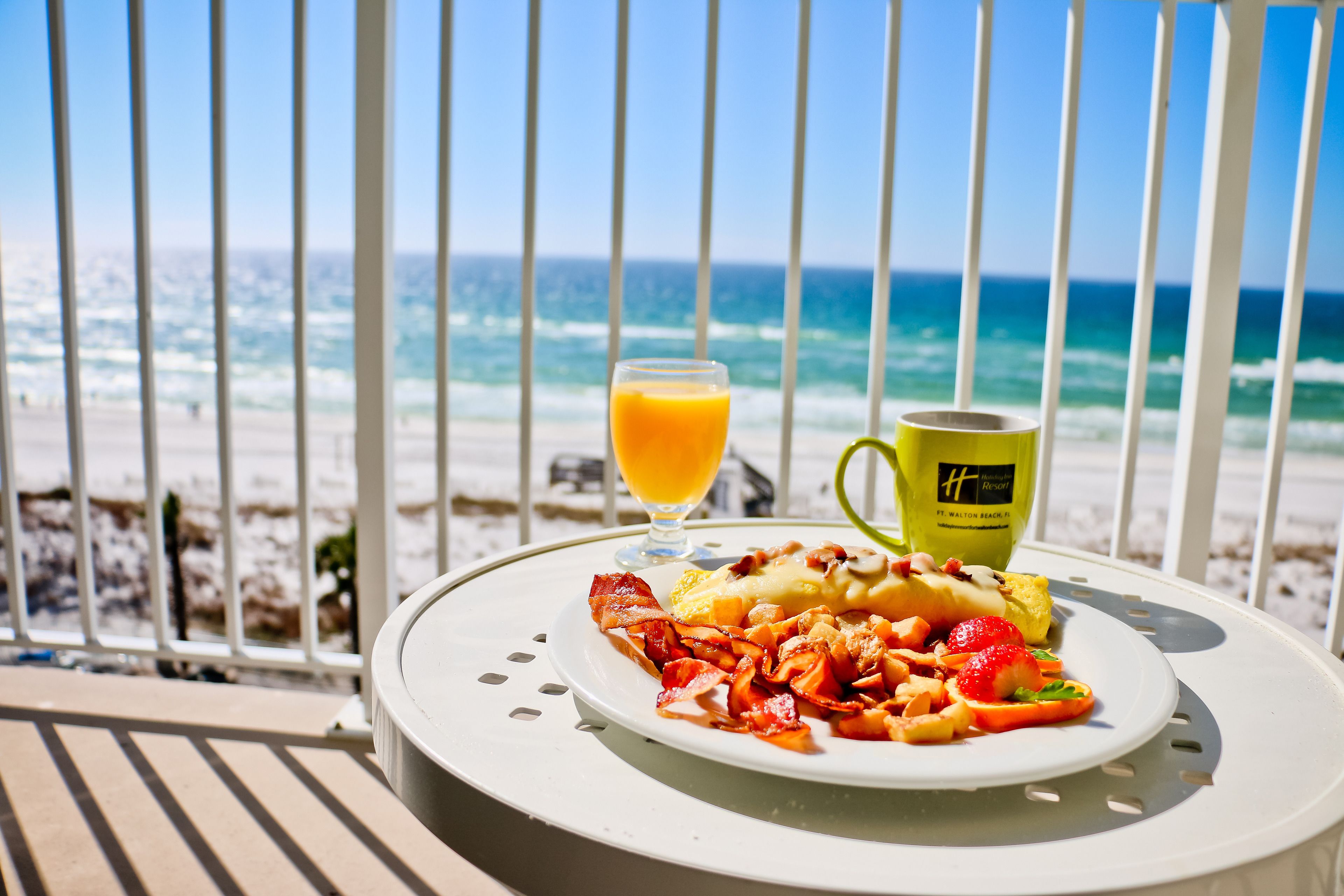 Une table de petit-déjeuner avec une assiette de nourriture, un verre de jus d’orange et une tasse, donnant sur une plage avec l’océan en arrière-plan.