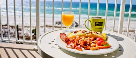 A breakfast table with a plate of food, a glass of orange juice, and a mug, overlooking a beach with the ocean in the background.