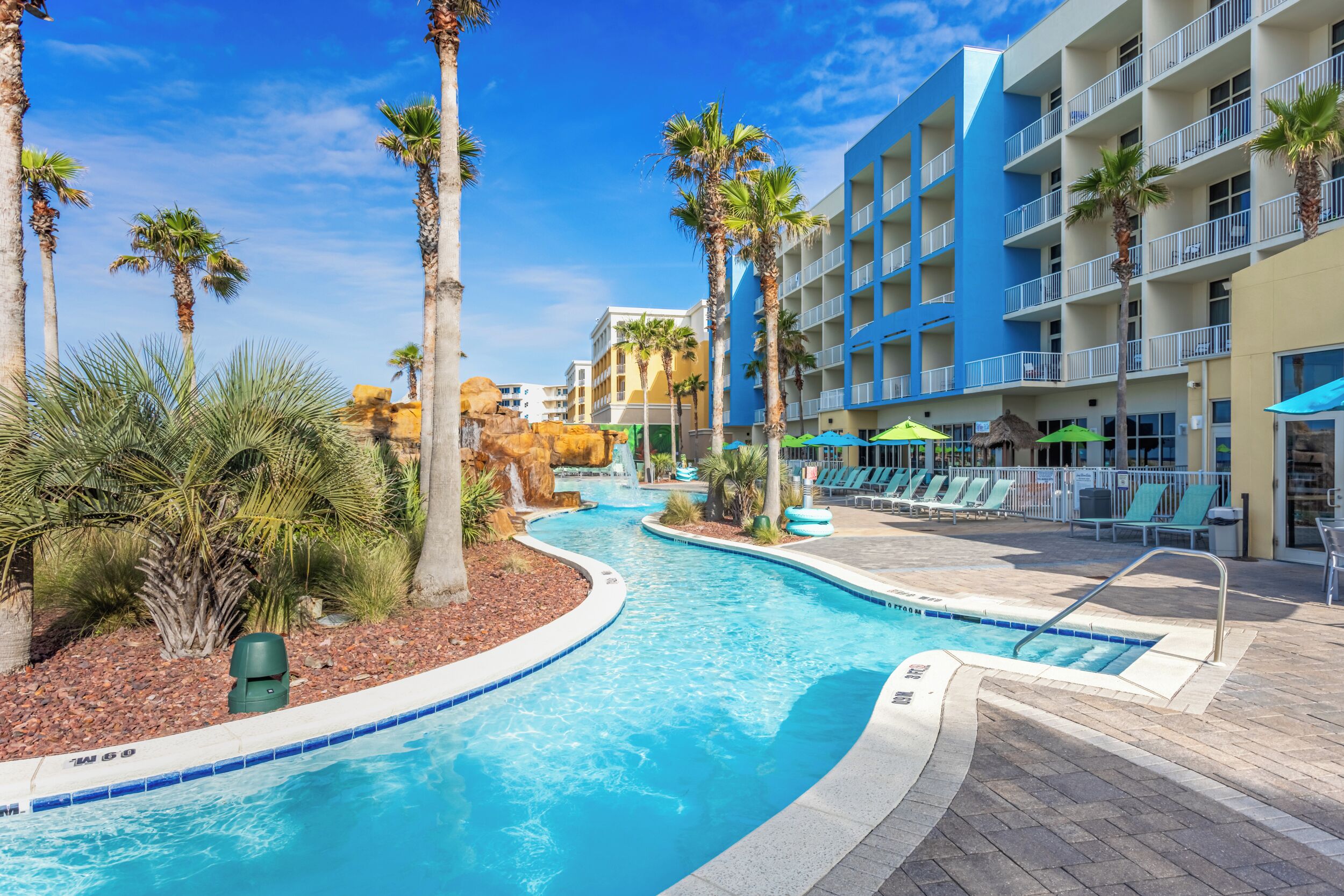 A swimming pool with palm trees and lounge chairs, surrounded by a hotel building with balconies.