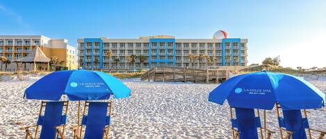 A beach with blue umbrellas and lounge chairs, a hotel building with balconies, and palm trees.