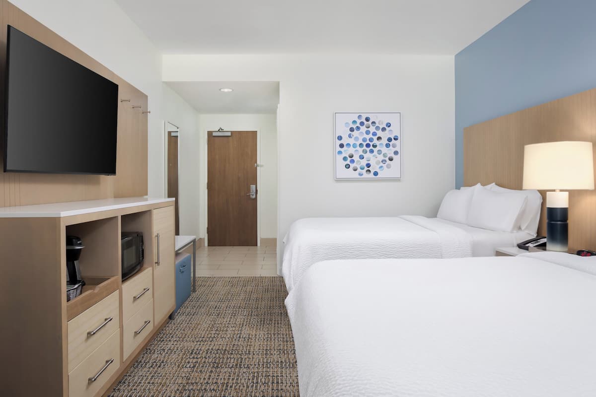 A hotel room with two beds, a flat-screen TV, a microwave, a desk, a lamp, and a wall art piece.