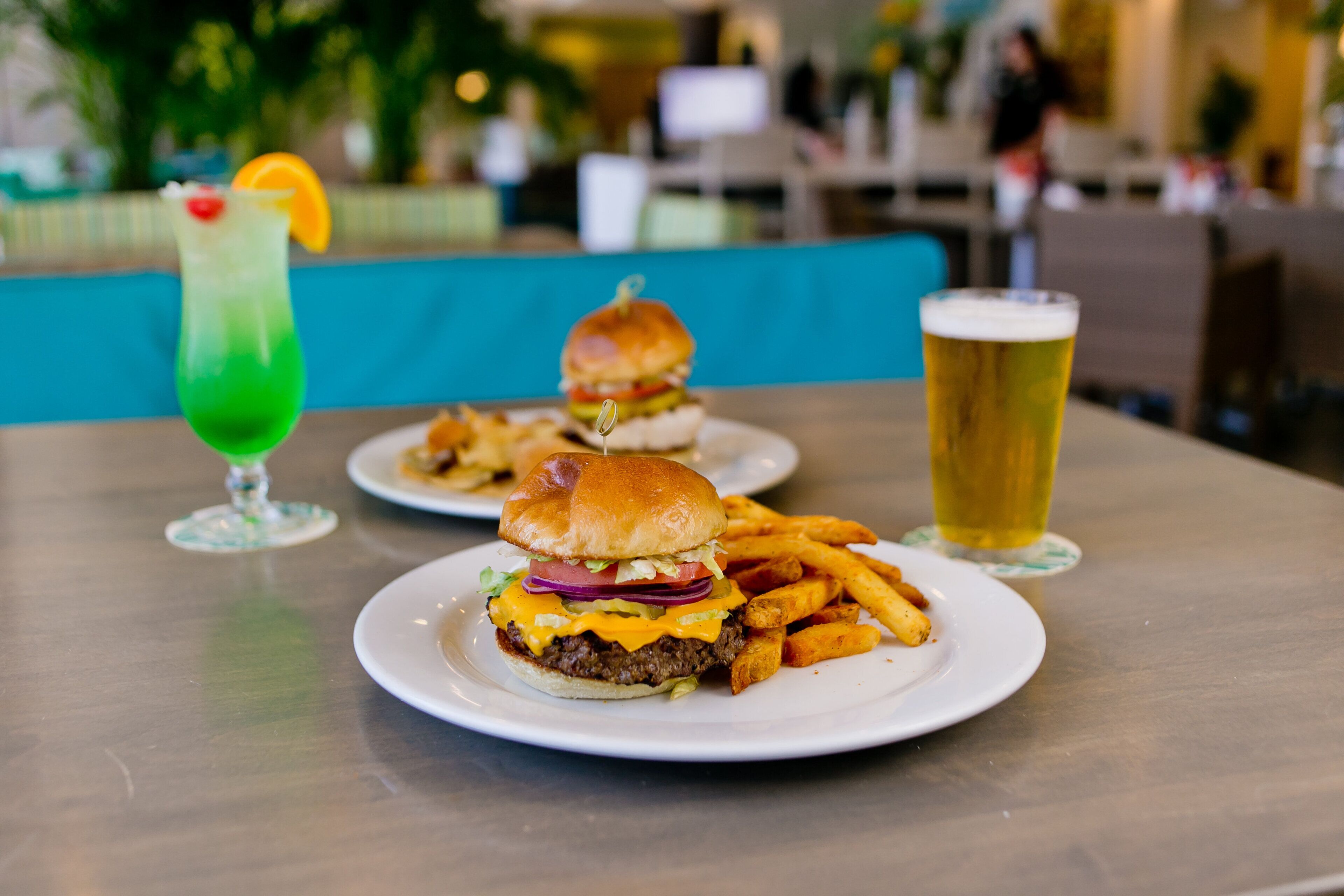 Un ambiente de restaurante con una hamburguesa y papas fritas sobre una mesa, una bebida verde y una jarra de cerveza.