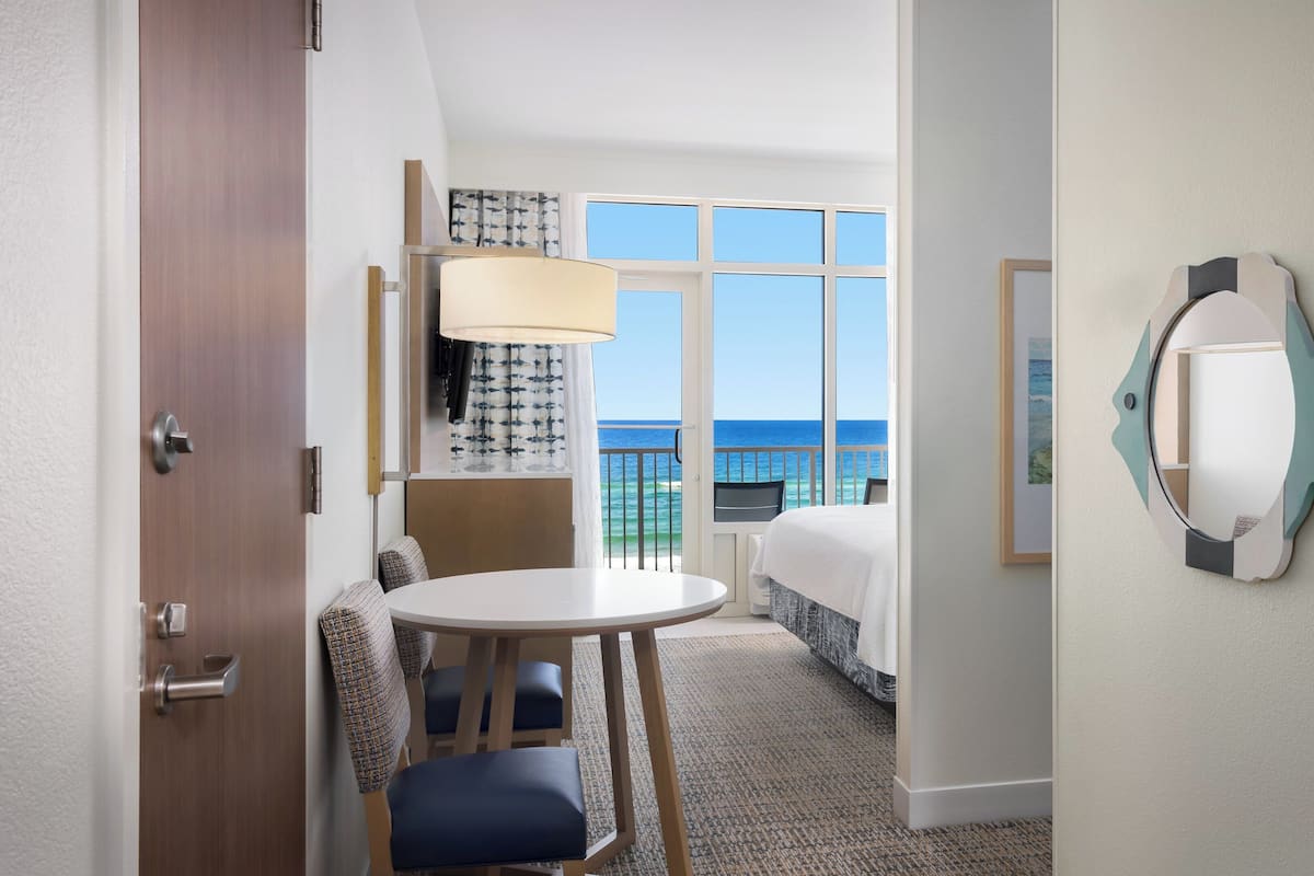 A hotel room with a bed, a round table, a chair, a window with a view of the sea, and a decorative mirror.