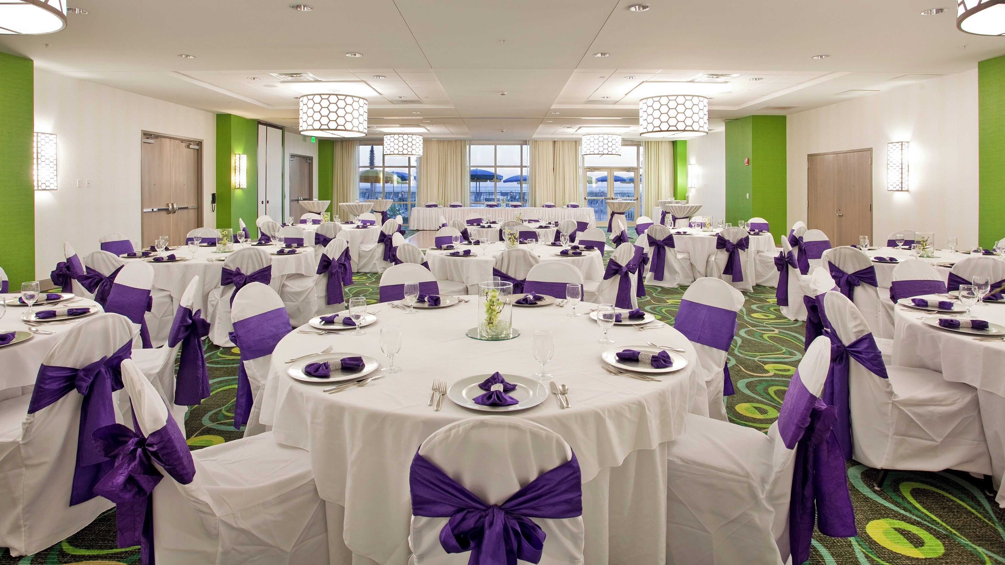 A banquet hall with round tables, chairs with purple sashes, and a view of the outdoors through large windows.