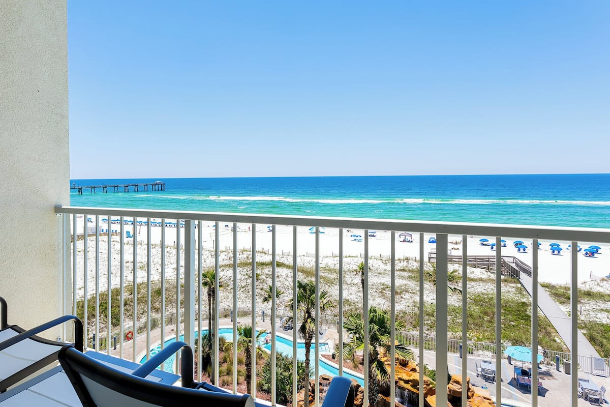 A balcony with a view of a beach, a pool, and a pier.
