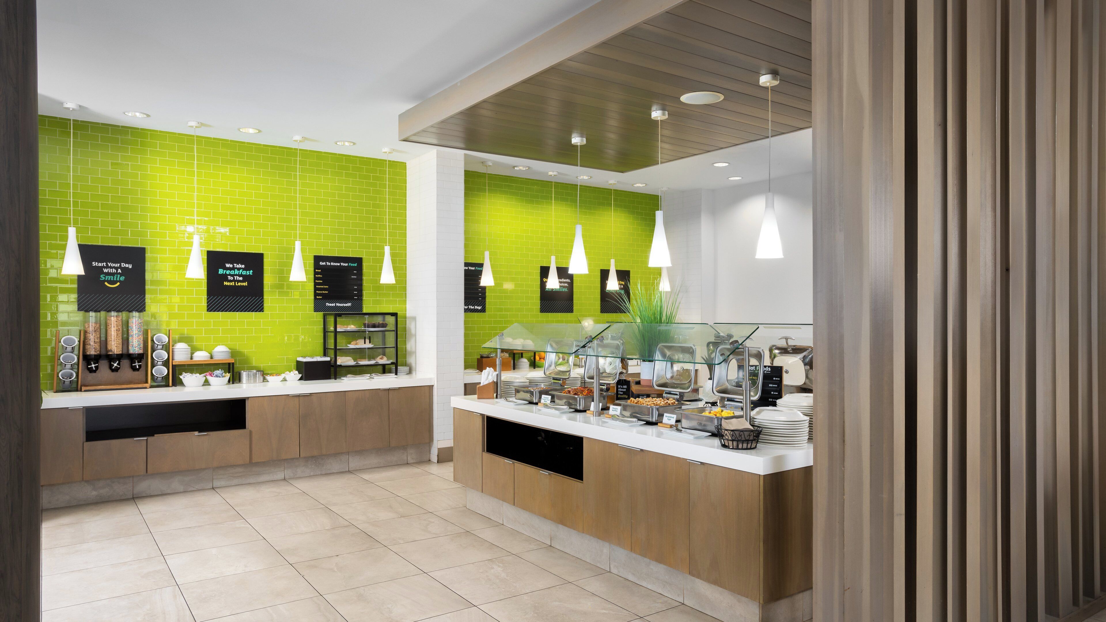 A modern breakfast area with a green-tiled wall, wooden cabinets, and a glass display case filled with food items.