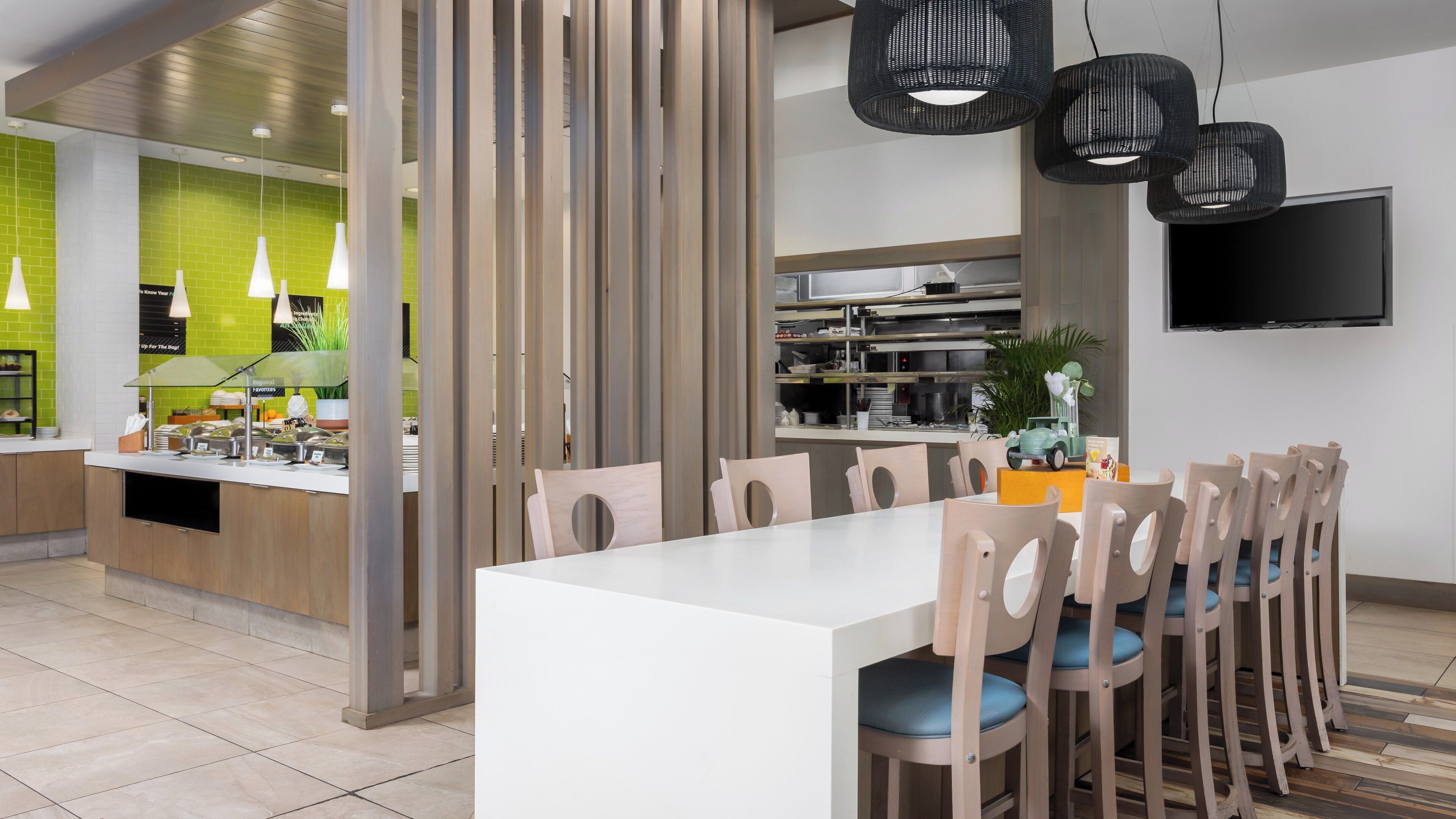 A modern dining area with a white table and wooden chairs, a kitchen in the background, and a TV mounted on the wall.