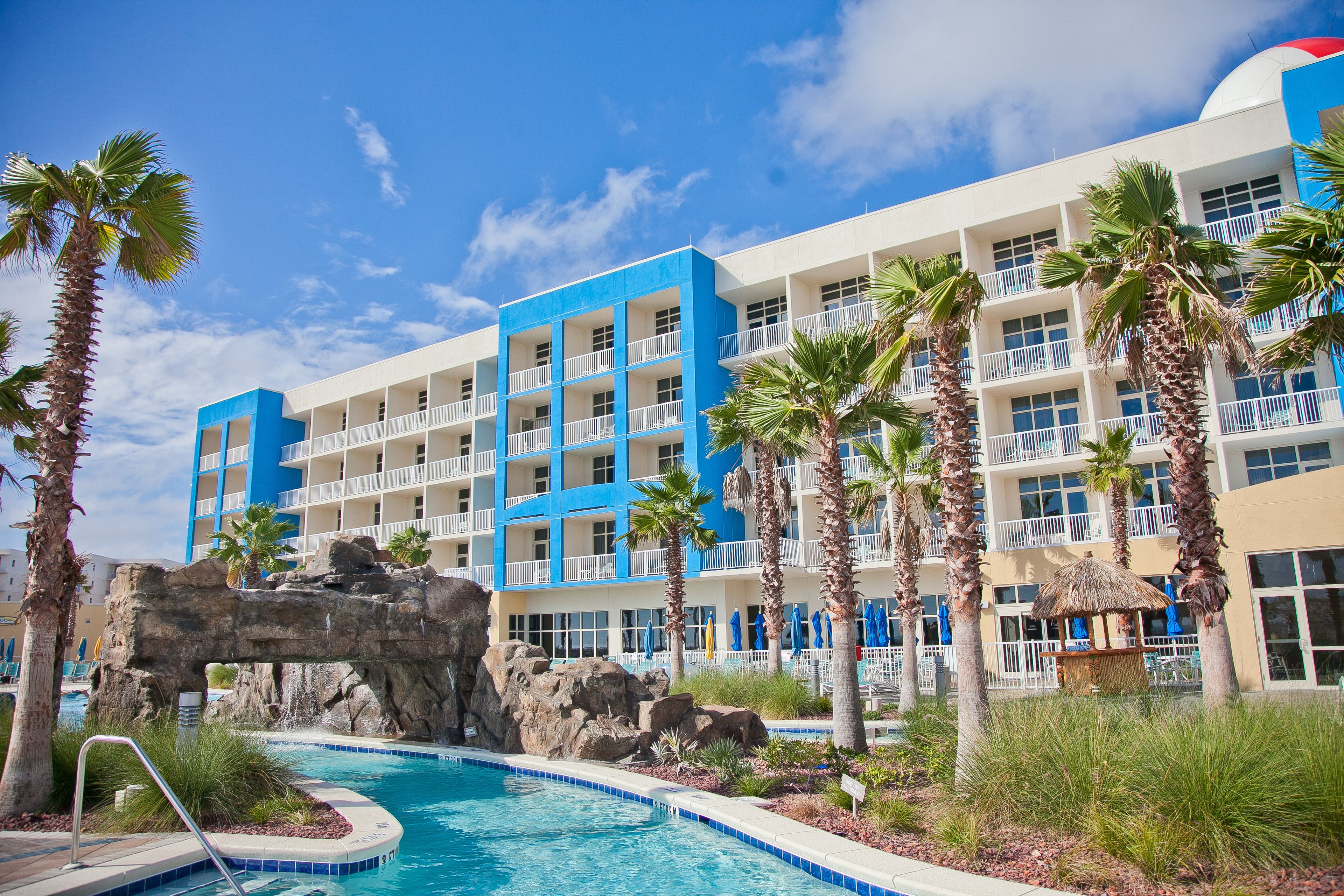 A hotel with multiple balconies, a swimming pool with a rock feature, and palm trees.