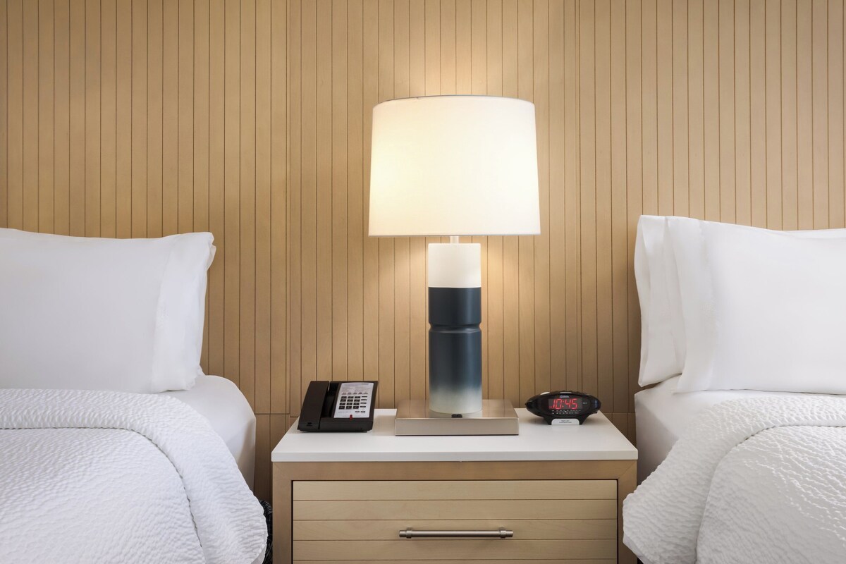A hotel room with two beds, a nightstand with a lamp, a phone, and a digital clock.