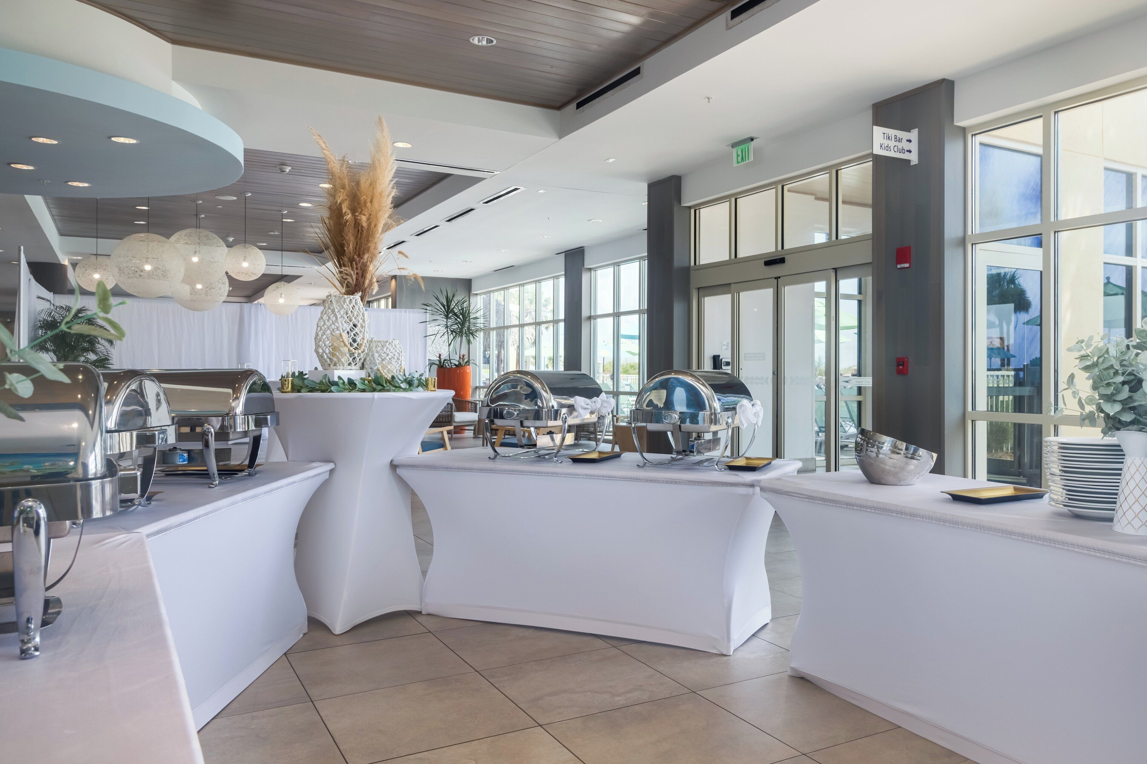 A modern, well-lit buffet area with stainless steel appliances and white tables, featuring large windows and natural light.