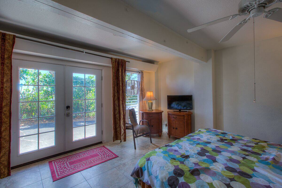 A bedroom with a bed, a television, a desk with a lamp, and a chair. There is a sliding glass door leading to a balcony.