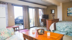 A hotel room with a bed, a sofa, a wooden table, a television, and a view of the outside. - Kona Hawaii Guesthouse (Kailua-Kona)