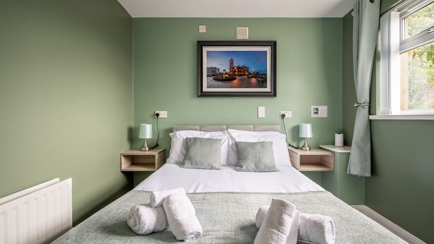 A neatly made bed with white linens and pillows, a framed picture of a cityscape on the wall, and a window with curtains.