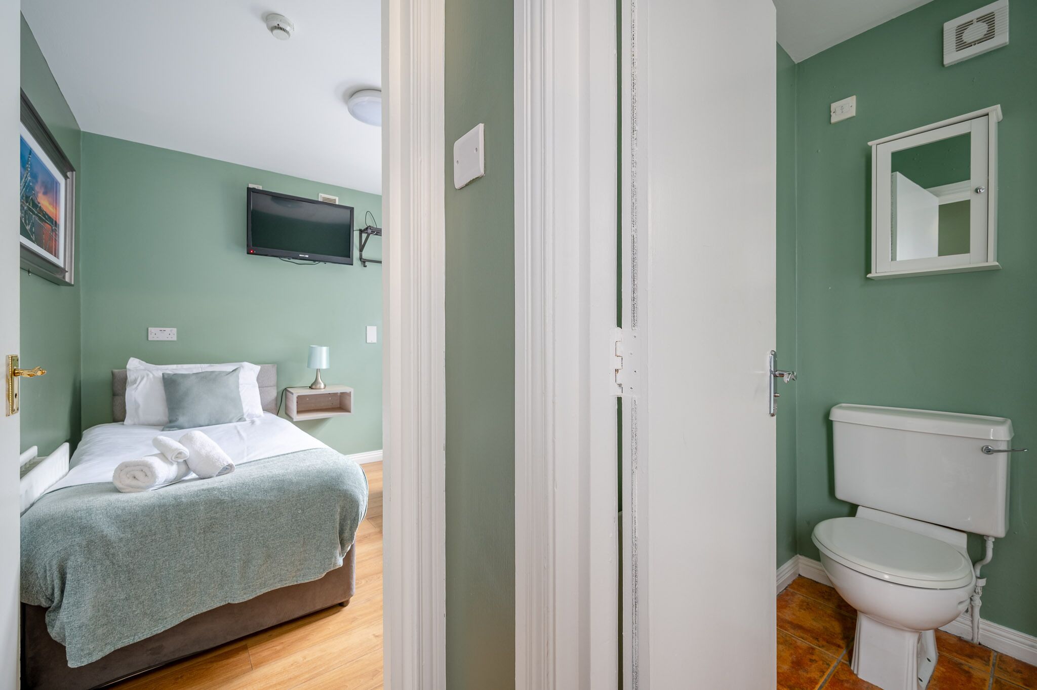 A bedroom with a bed, a bedside table with a lamp, a television on the wall, and a bathroom with a toilet and sink.