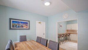A dining area with a wooden table and chairs, a painting on the wall, and a kitchen area with bar stools.