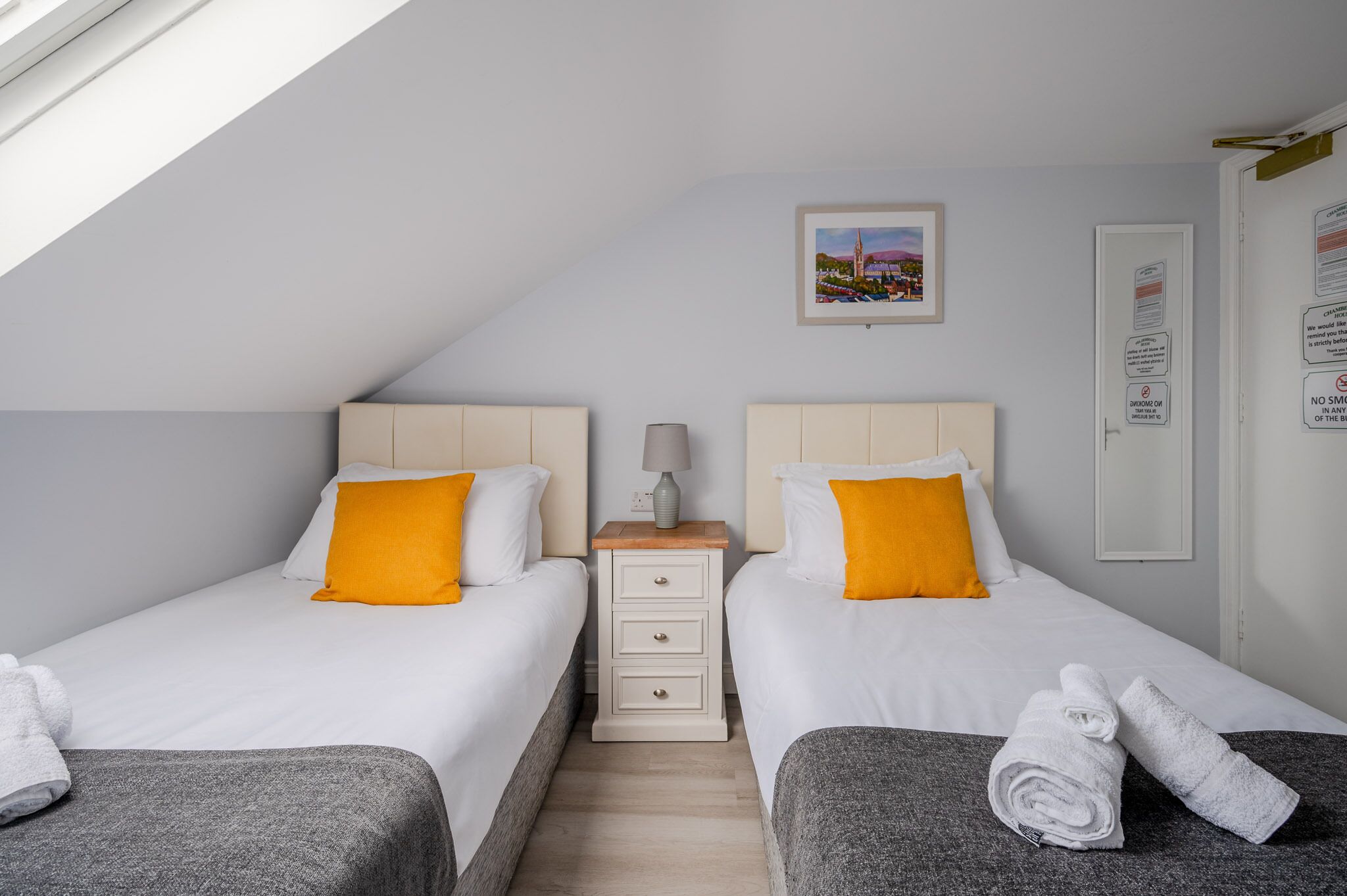 A hotel room with two beds, each with white bedding and orange pillows, a small bedside table with a lamp, and a picture on the wall.